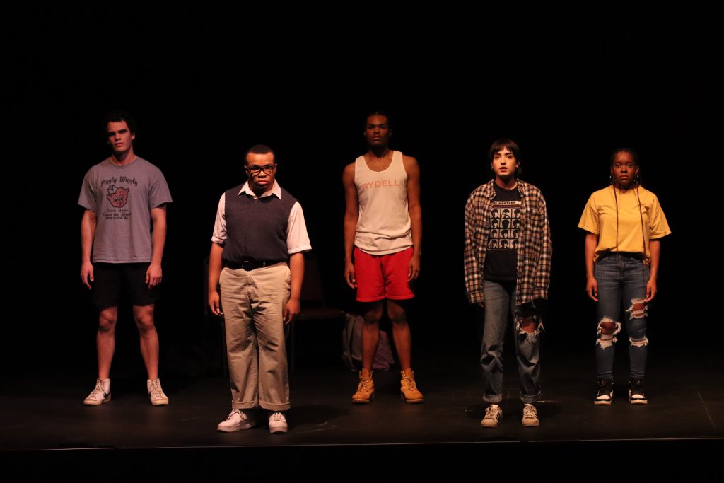 Five performers, standing in a line, make direct eye contact with the camera lens.