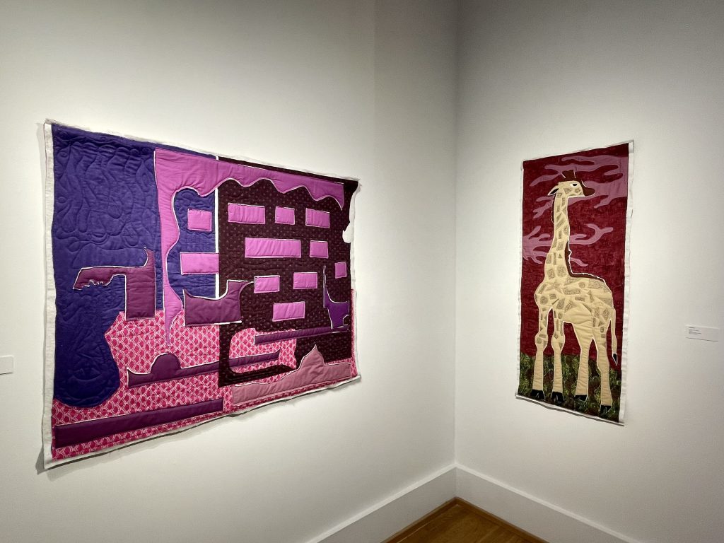 Two quilts, one with pink abstract shapes and the other a giraffe, hang on a wall.