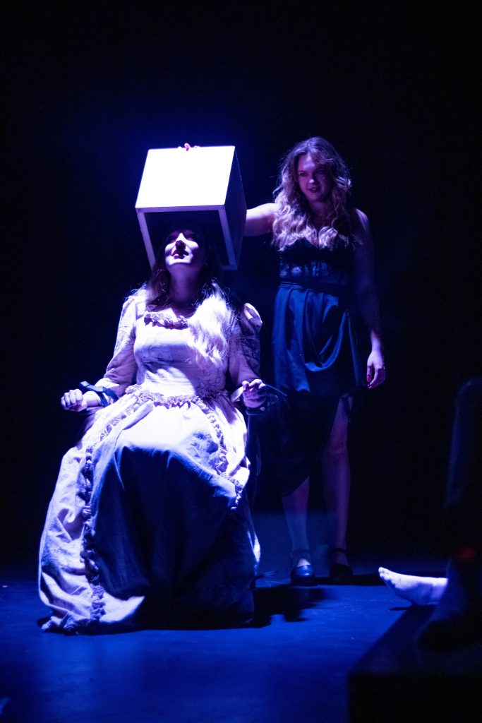 A woman sits tied to a chair while another places a box over her head.